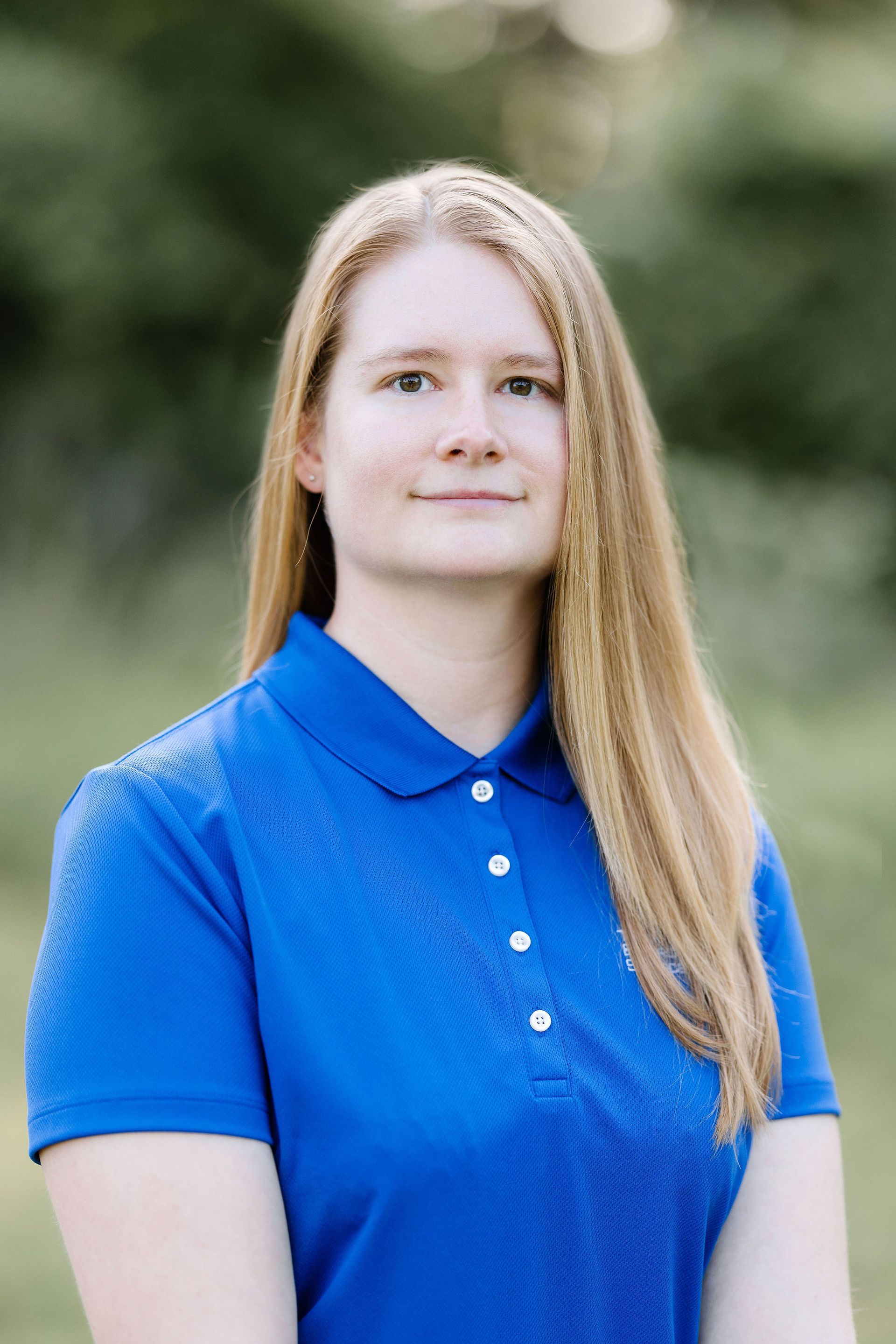 Woman with long blonde hair wearing a blue polo shirt, smiling slightly against a blurred green background.