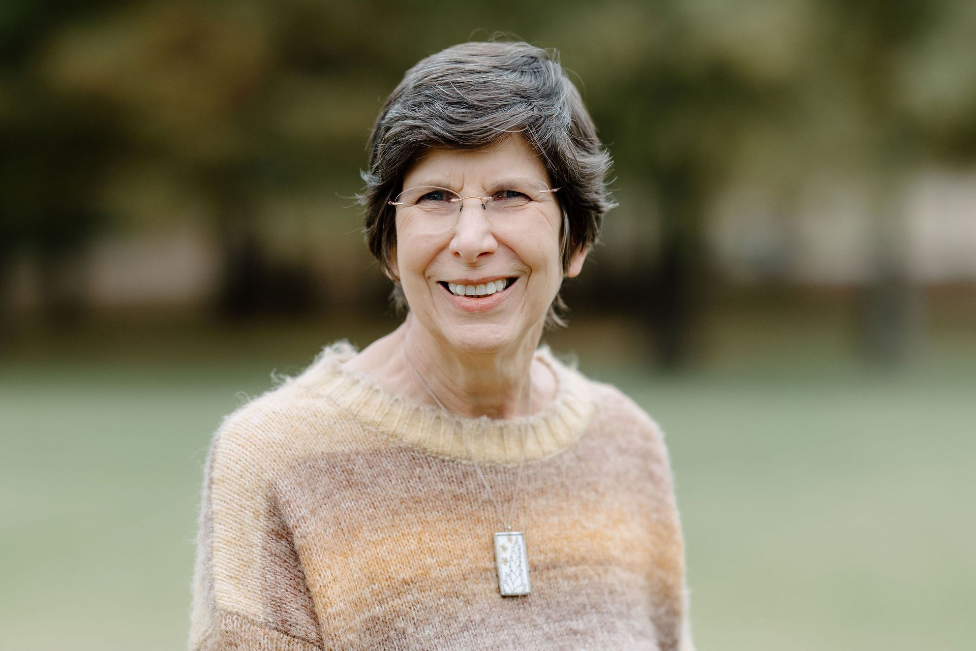 Woman with glasses smiles outdoors, wearing a tan and yellow sweater with a pendant.