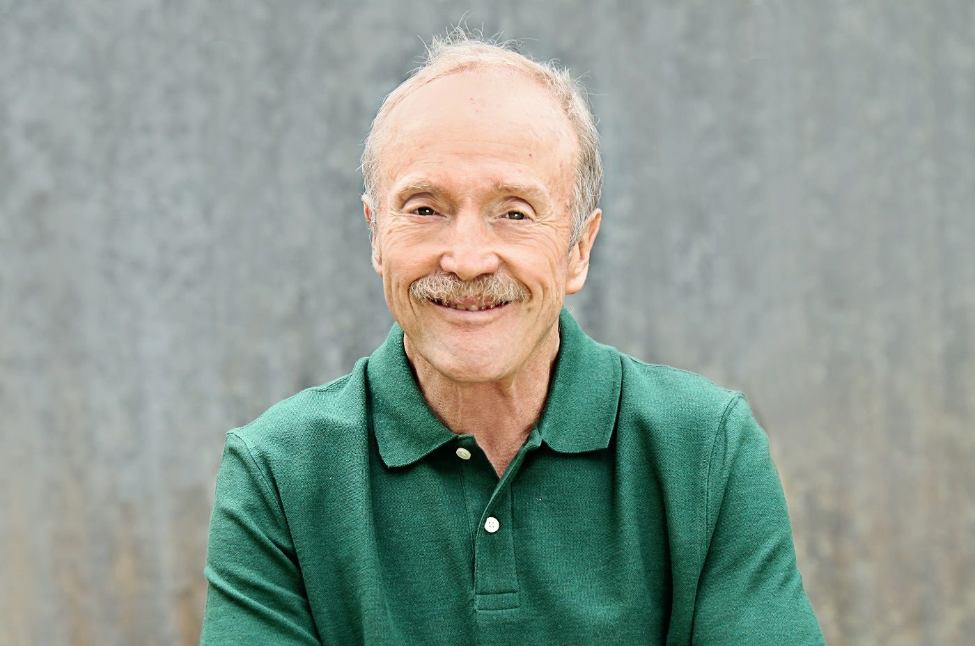 Man with a mustache wearing a green polo shirt, smiling in front of a gray backdrop.