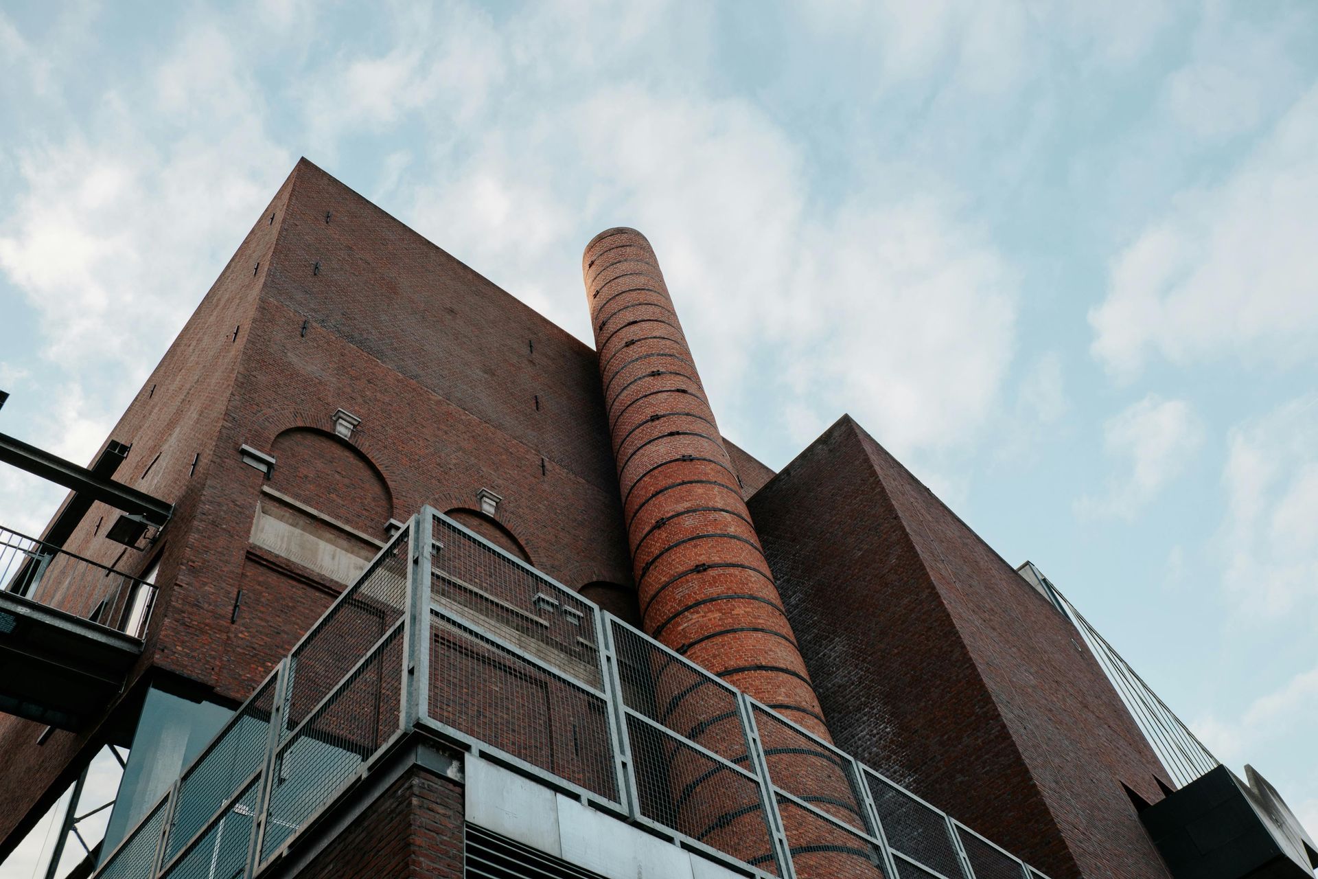 Red brick building with tall chimney, metal walkways, against a cloudy sky.