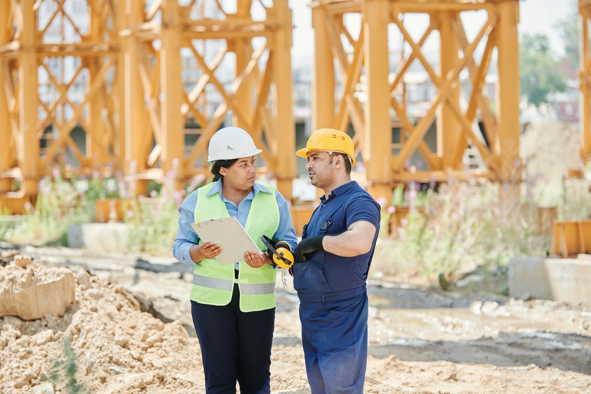 Two construction workers reviewing a clipboard at a building site with yellow cranes.