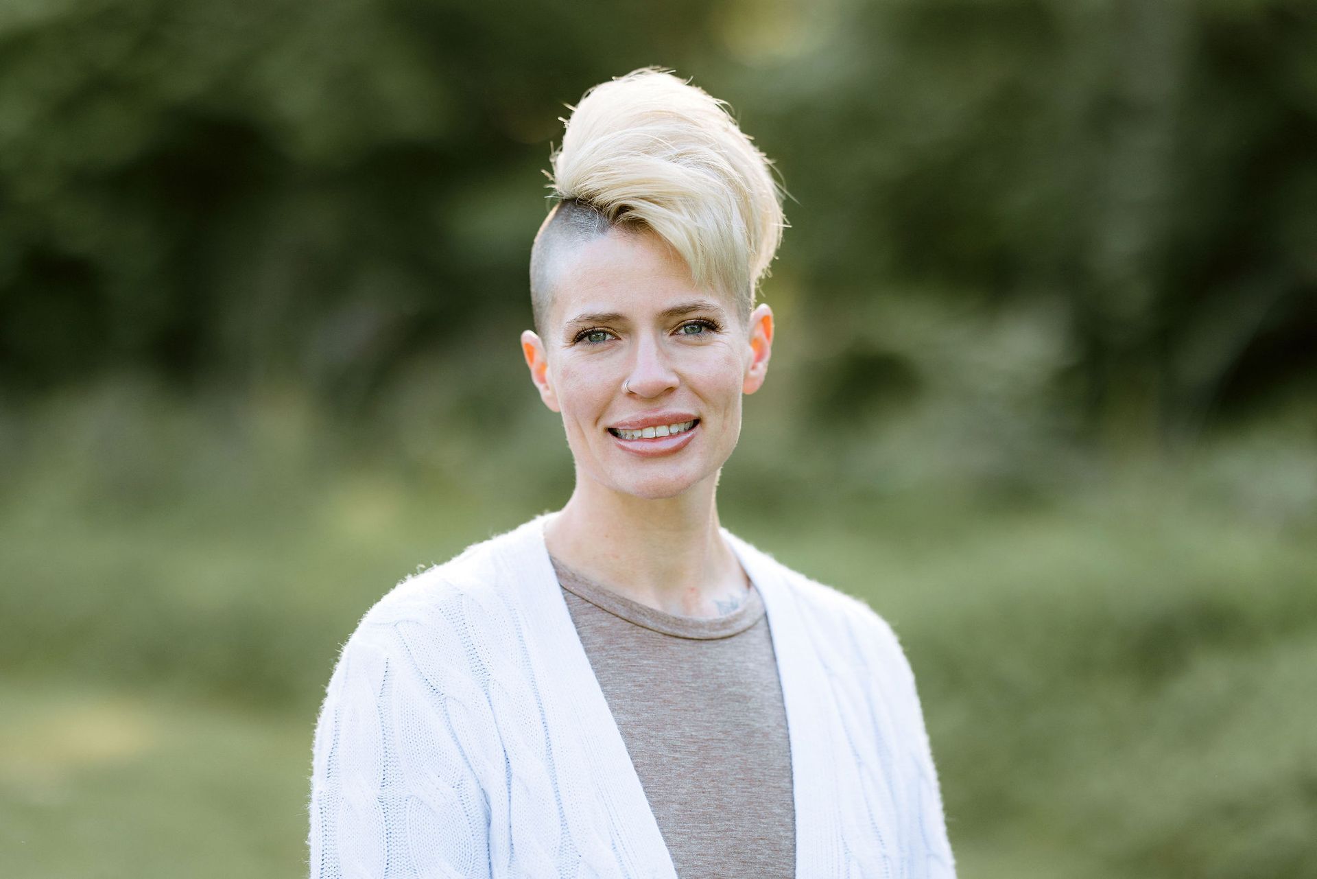 Woman with light blonde mohawk smiling, wearing a white cardigan and gray shirt, outdoors.