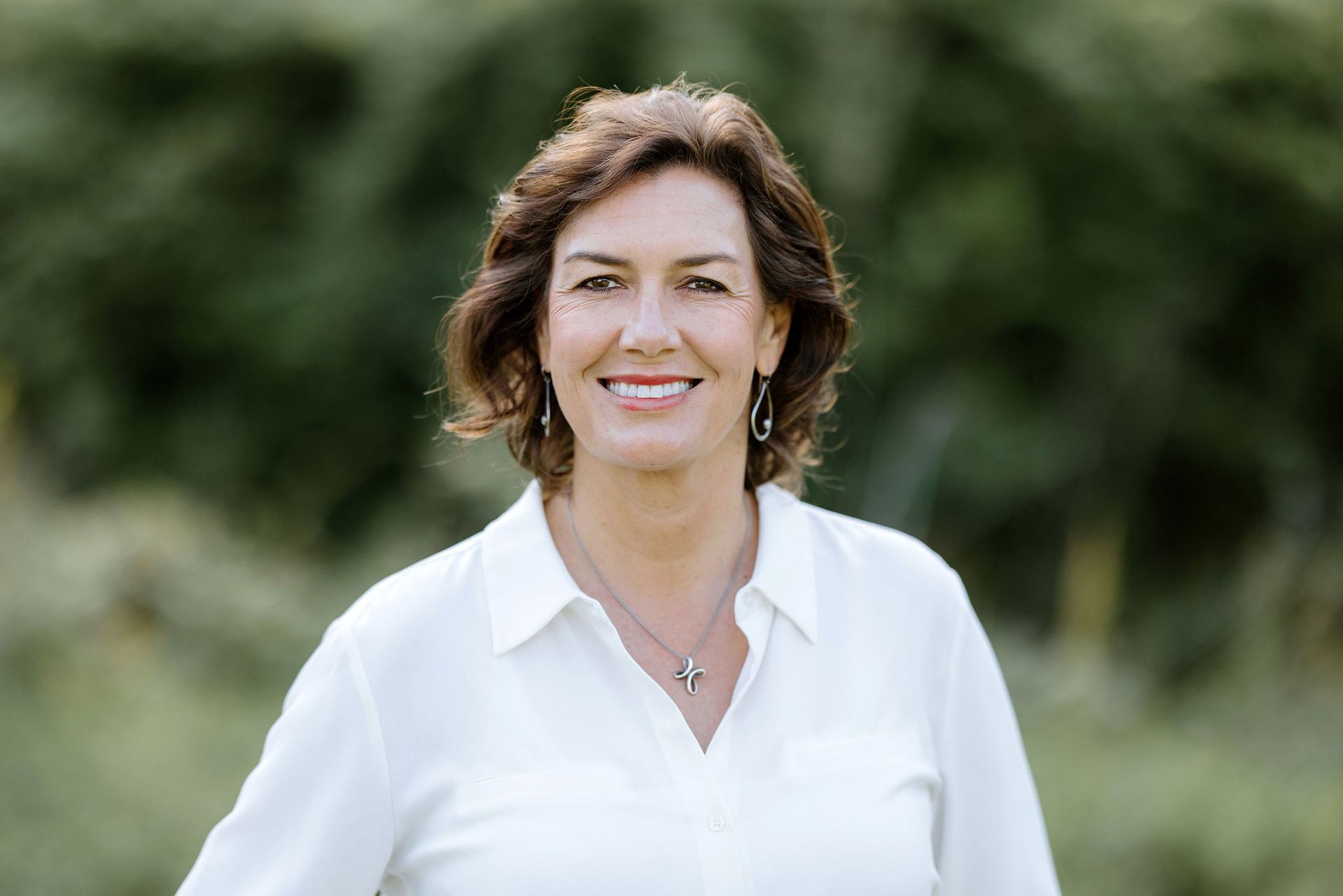 Woman in white shirt smiles outdoors with blurred green background.