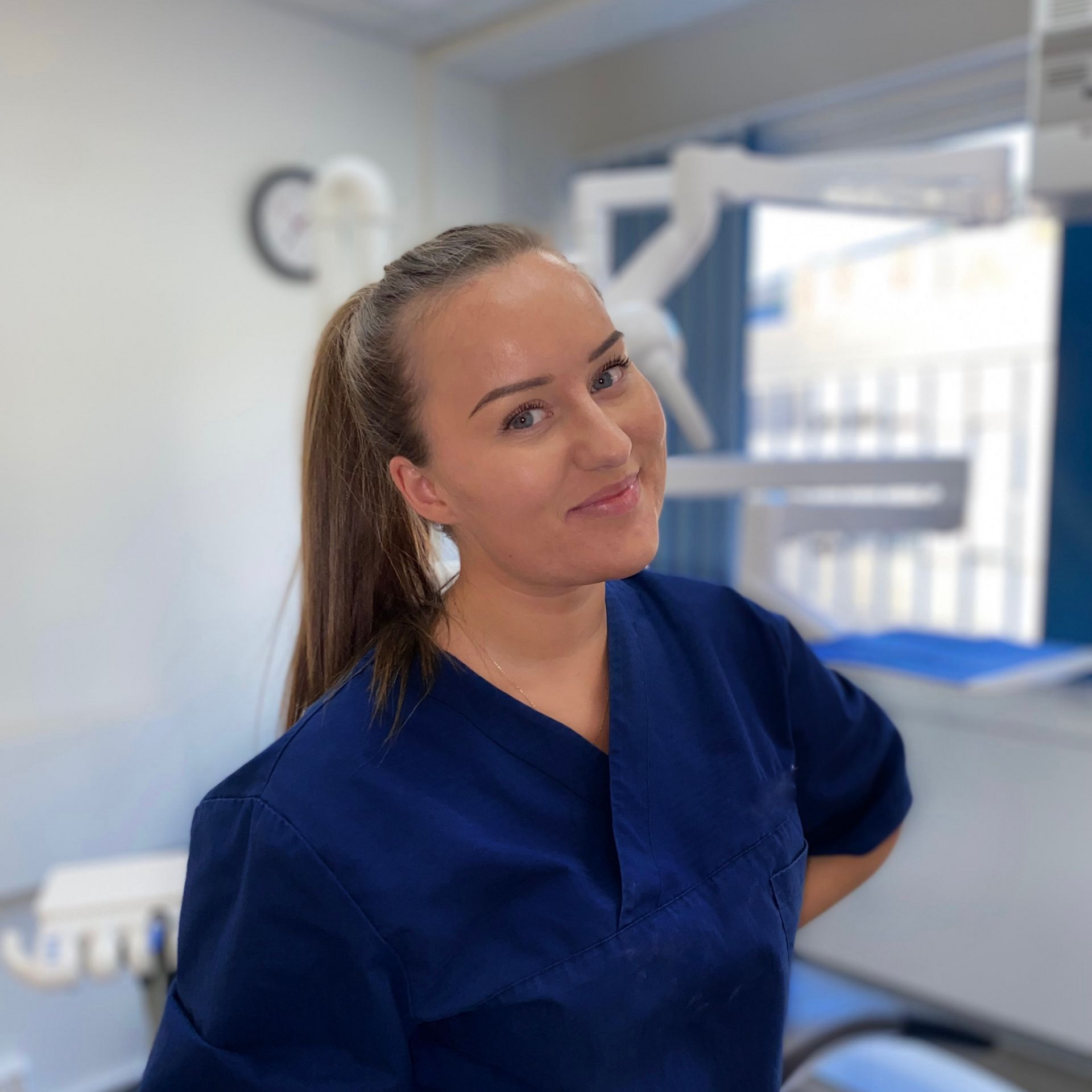 Kvinne med blondt hår smiler, iført en marineblå blazer og hvit skjorte, utendørs.