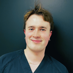 Tannlege Christian med briller og skjegg, iført blå skjorte og medisinsk maske, smiler til kameraet.