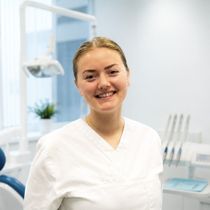 Kvinne med briller og blå skrubber smiler på et tannlegekontor.
