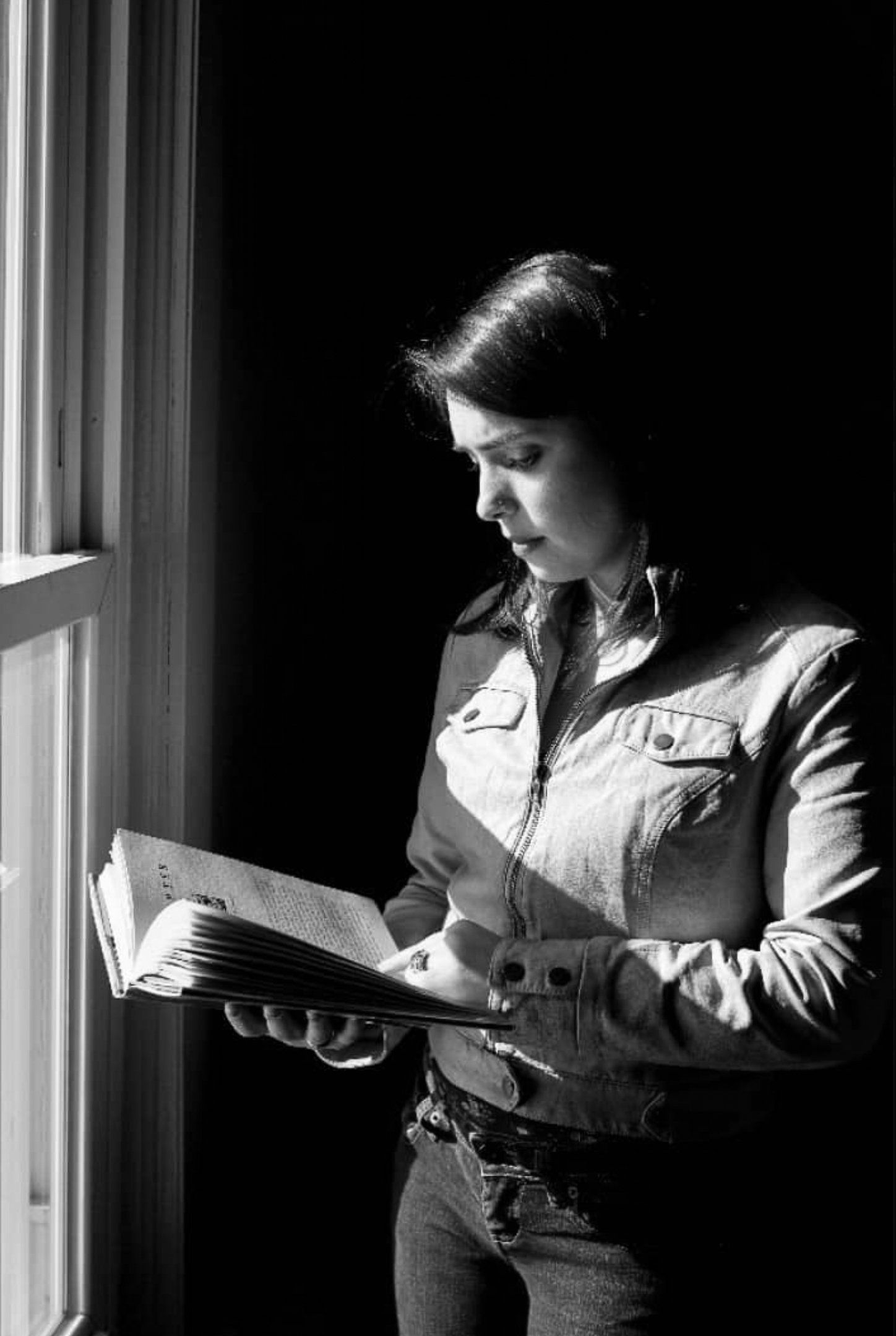 A woman is standing in front of a window reading a book.
