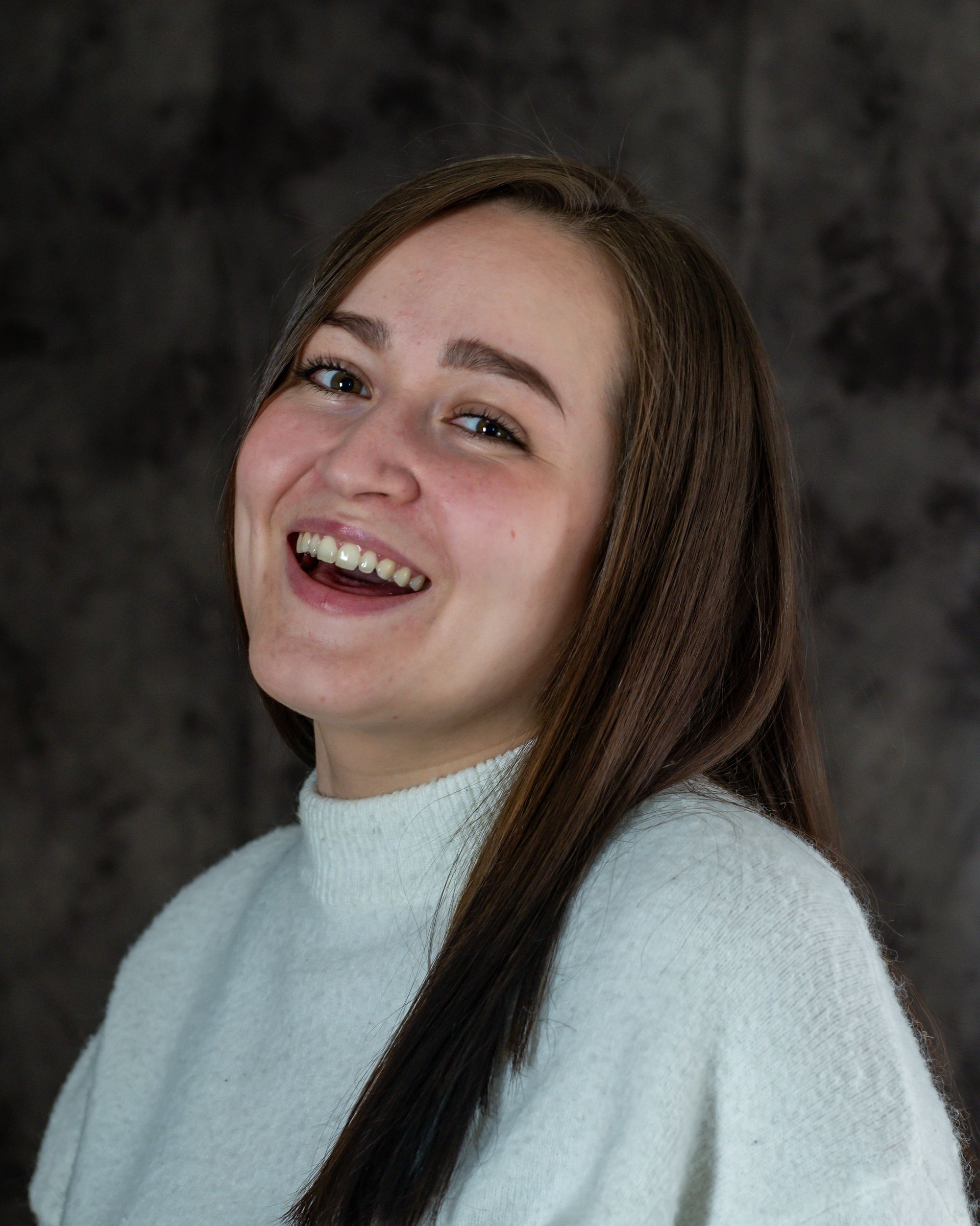 A woman with long brown hair is wearing a white sweater and smiling.