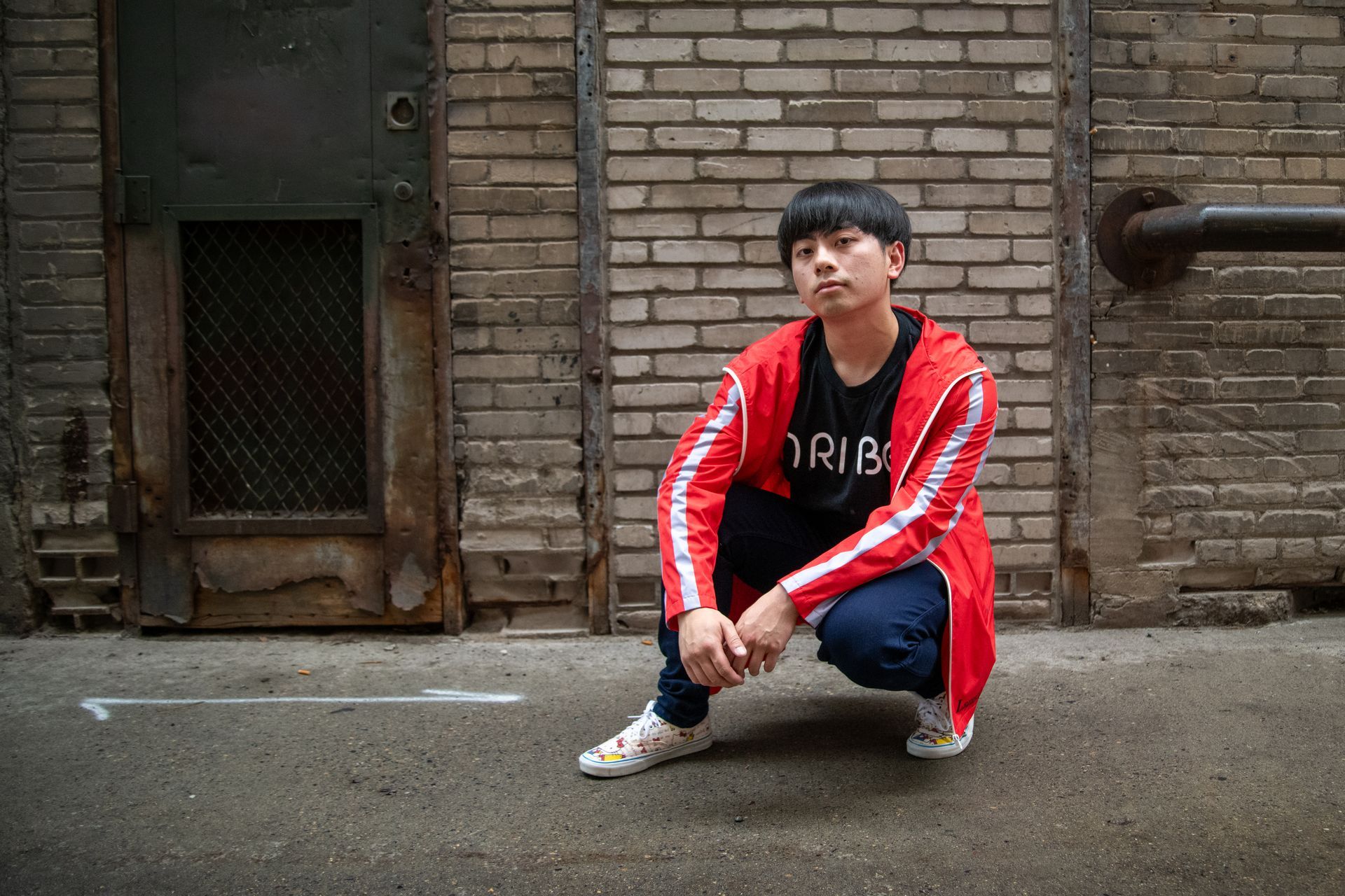 A man wearing a red jacket and a t-shirt that says trib is squatting in front of a brick wall