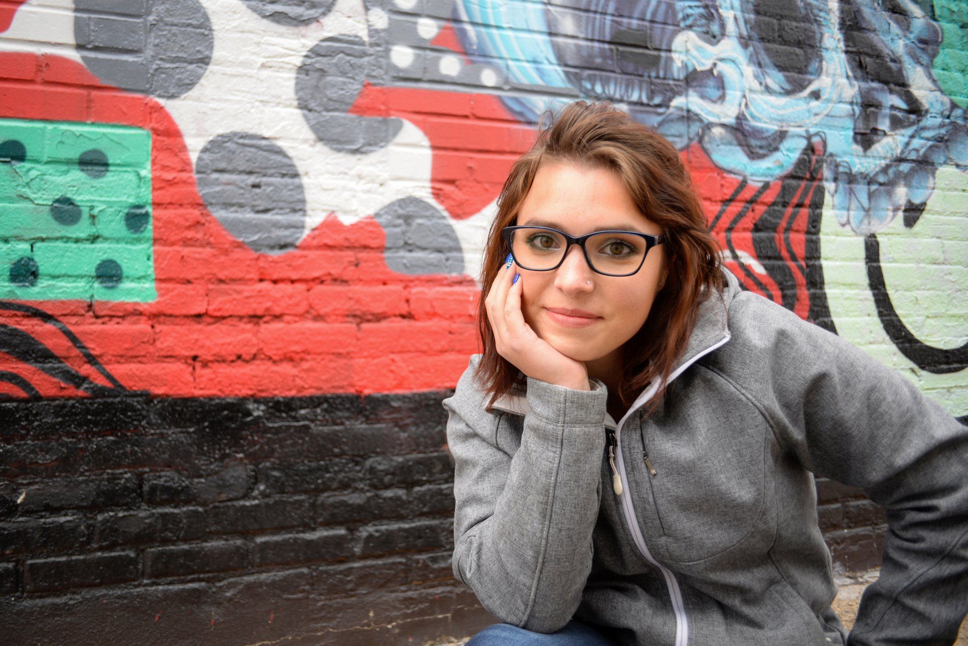 A woman wearing glasses is sitting in front of a brick wall with graffiti on it