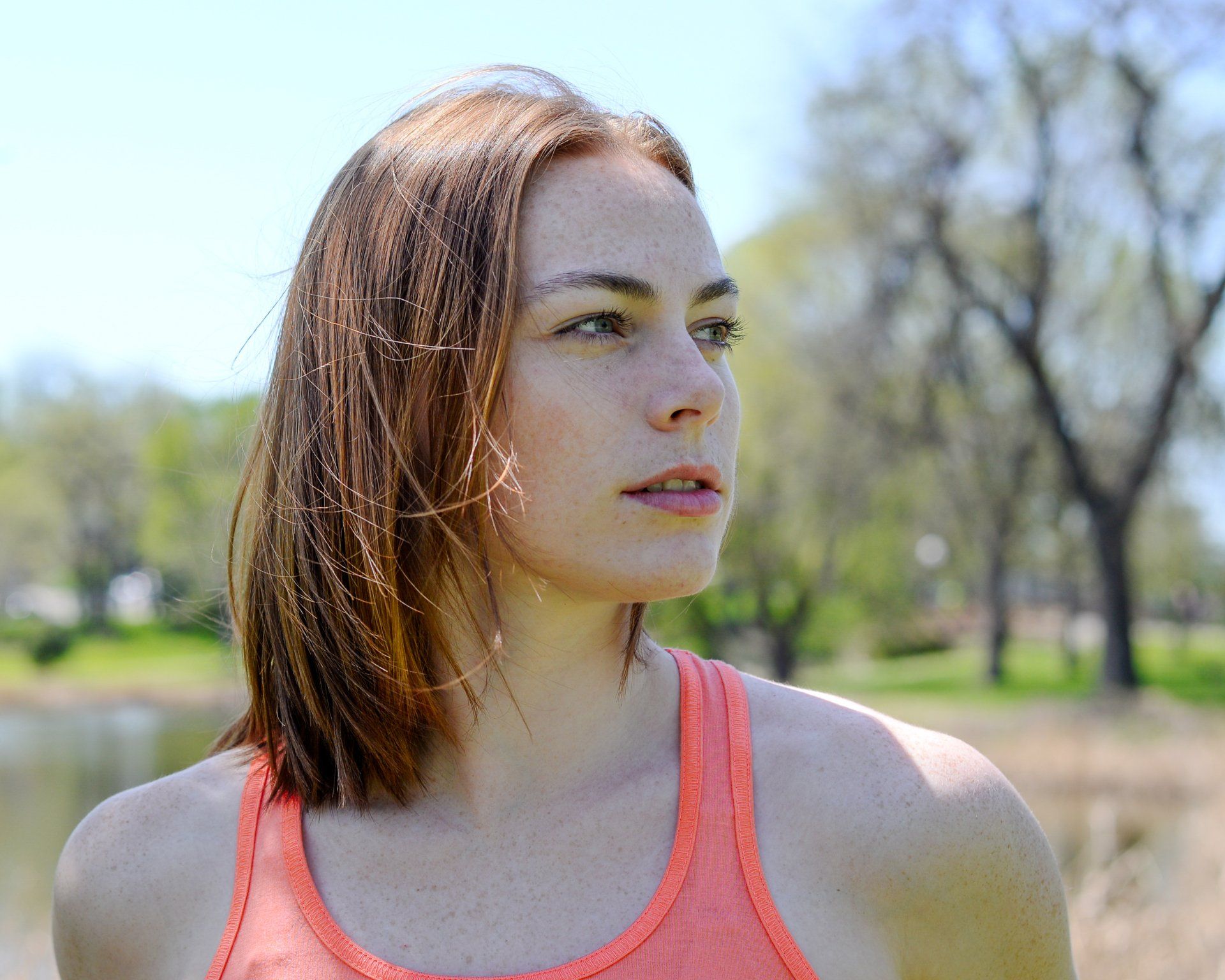 A woman in an orange tank top stands in front of a lake