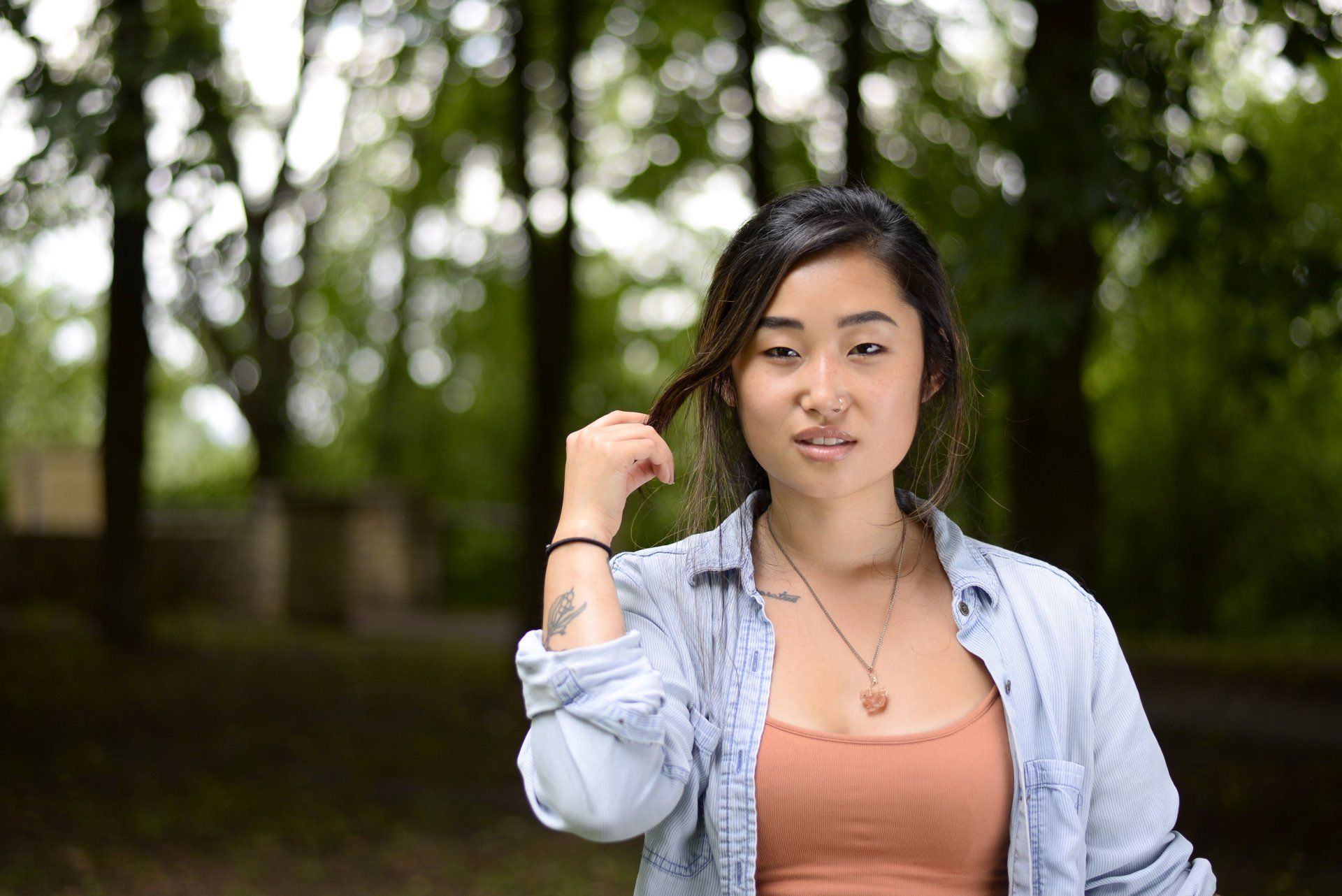 A woman in a denim shirt is holding her hair in front of trees