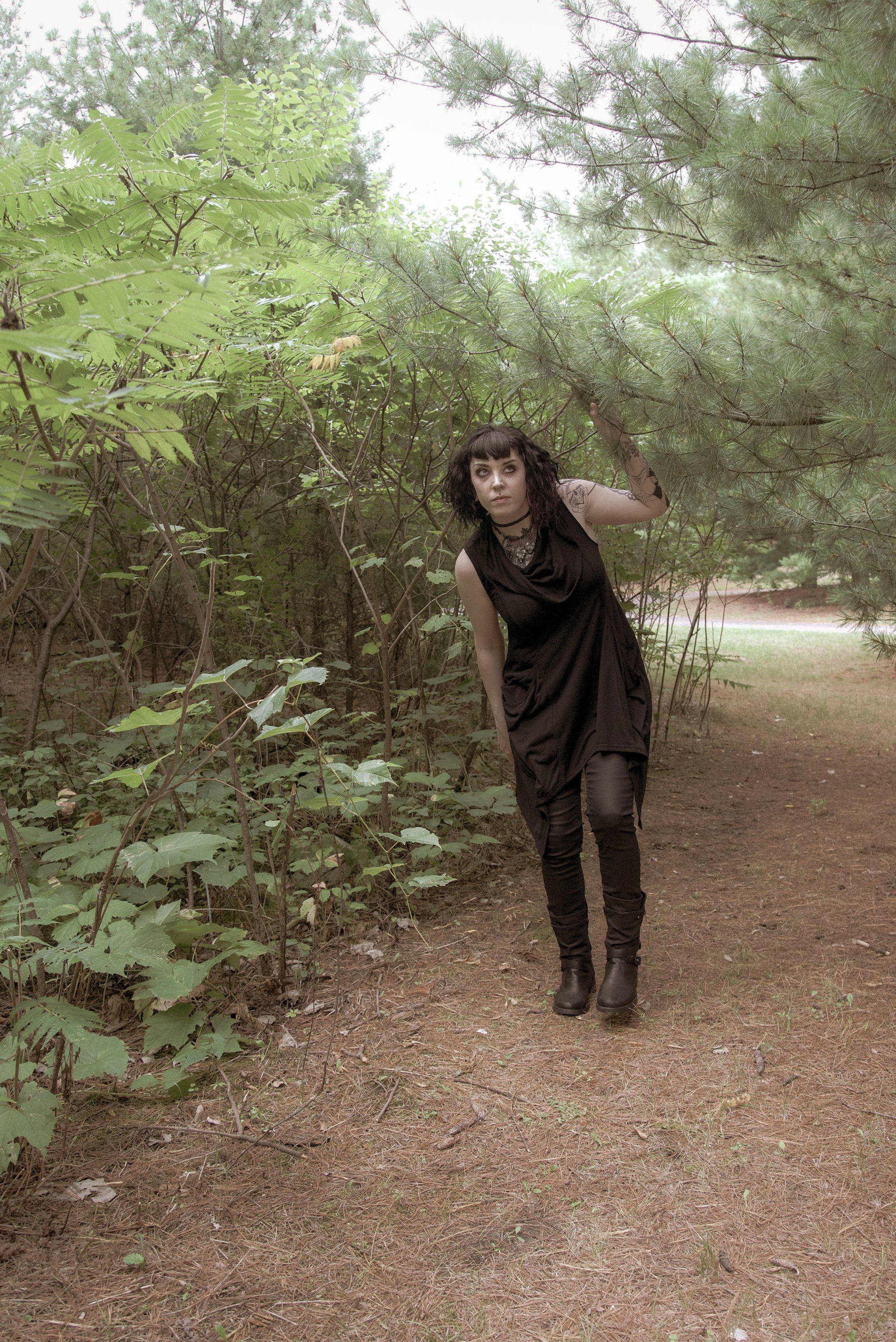 A woman in a black dress is standing on a dirt path in the woods.