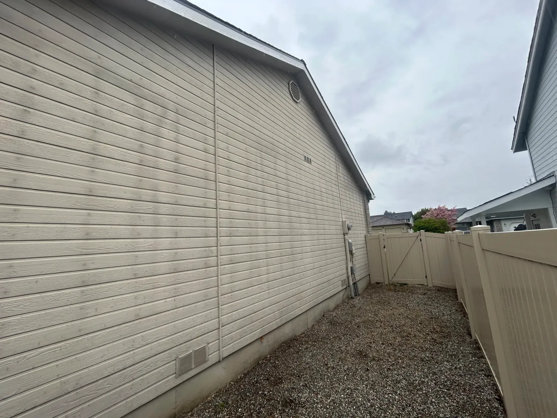 Side view of a house with beige siding, a gravel yard, and a tan fence under a cloudy sky.