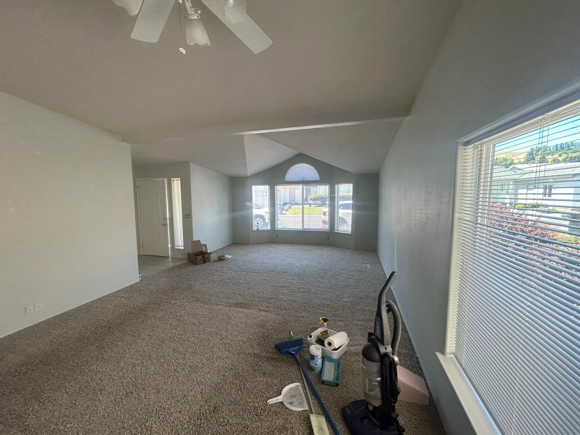 Empty living room with tan carpet, a large window, and cleaning supplies.