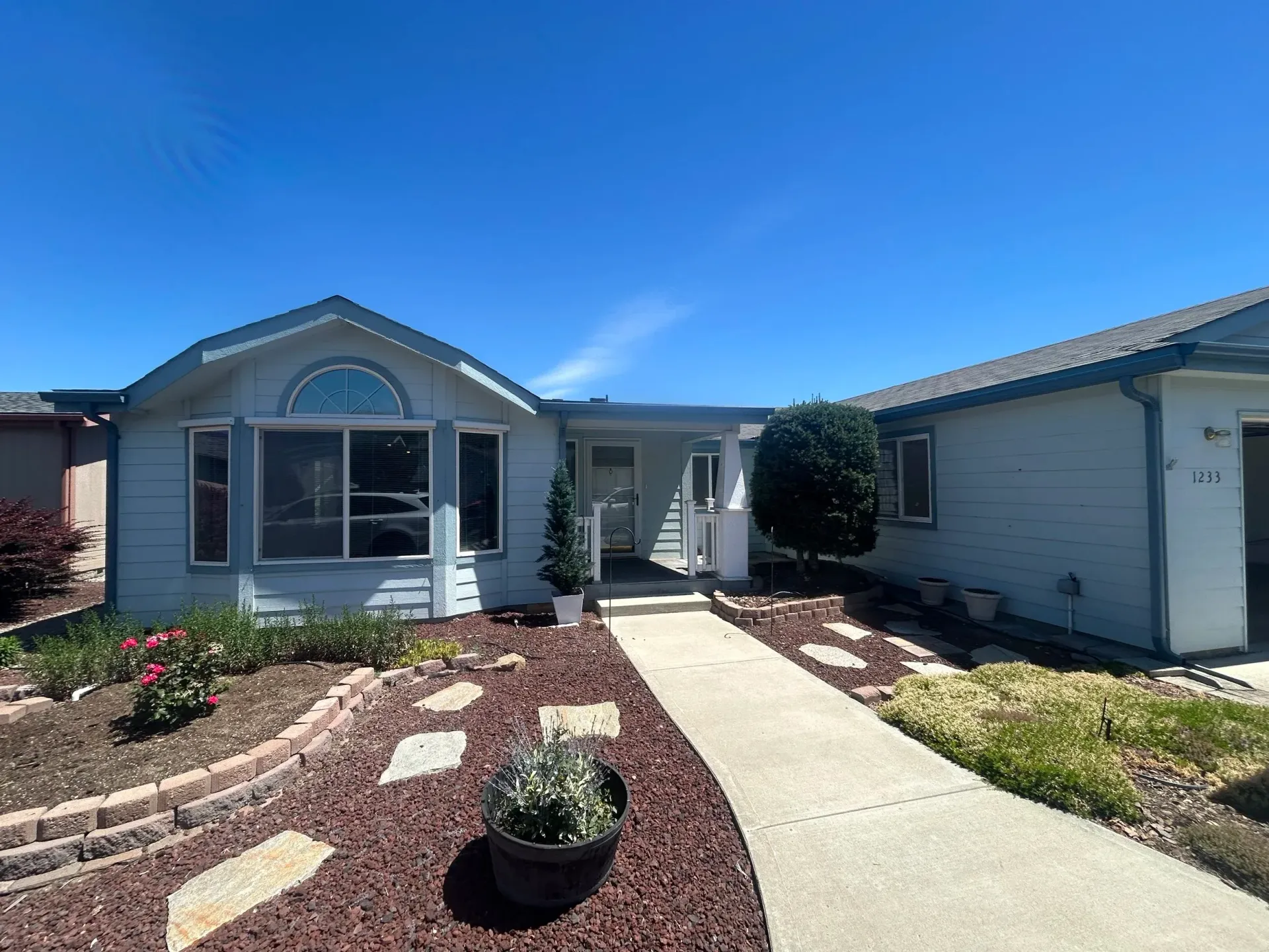 Front of a light blue house with a curved window, walkway, and small garden on a sunny day.