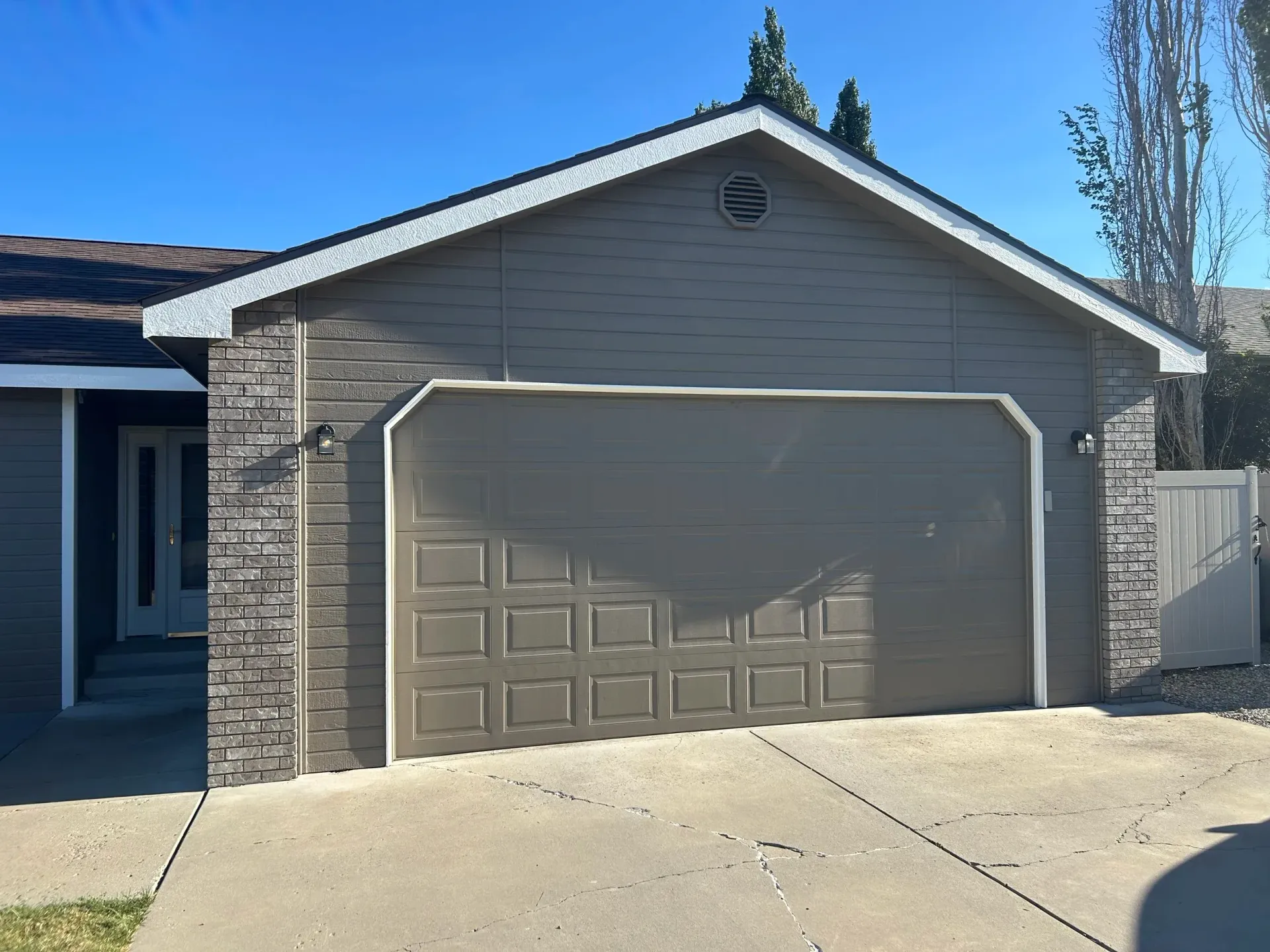 Garage with brown door and stone siding, gray house. Concrete driveway, clear sky.