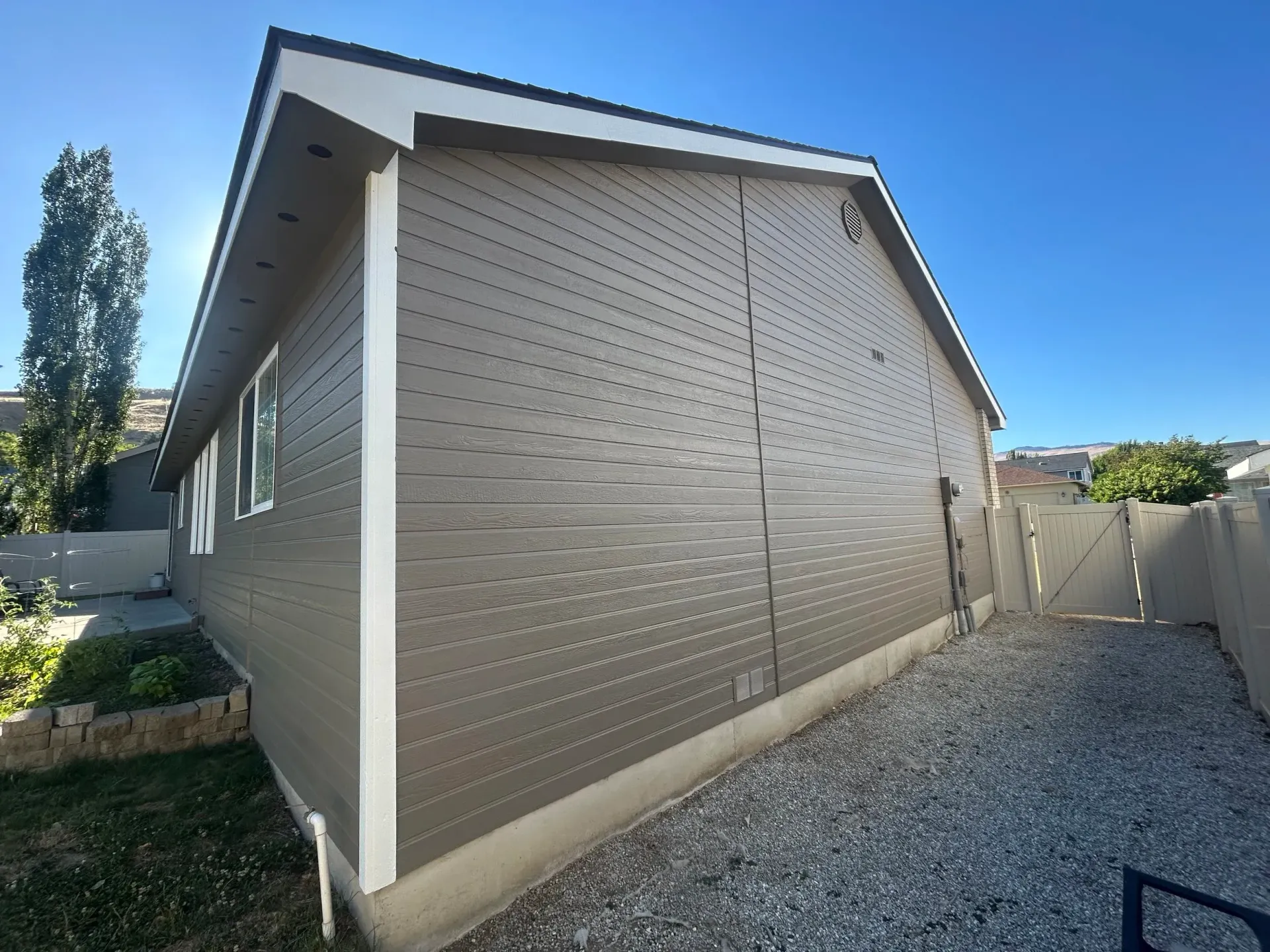 Tan sided house with white trim against a blue sky, gravel yard.