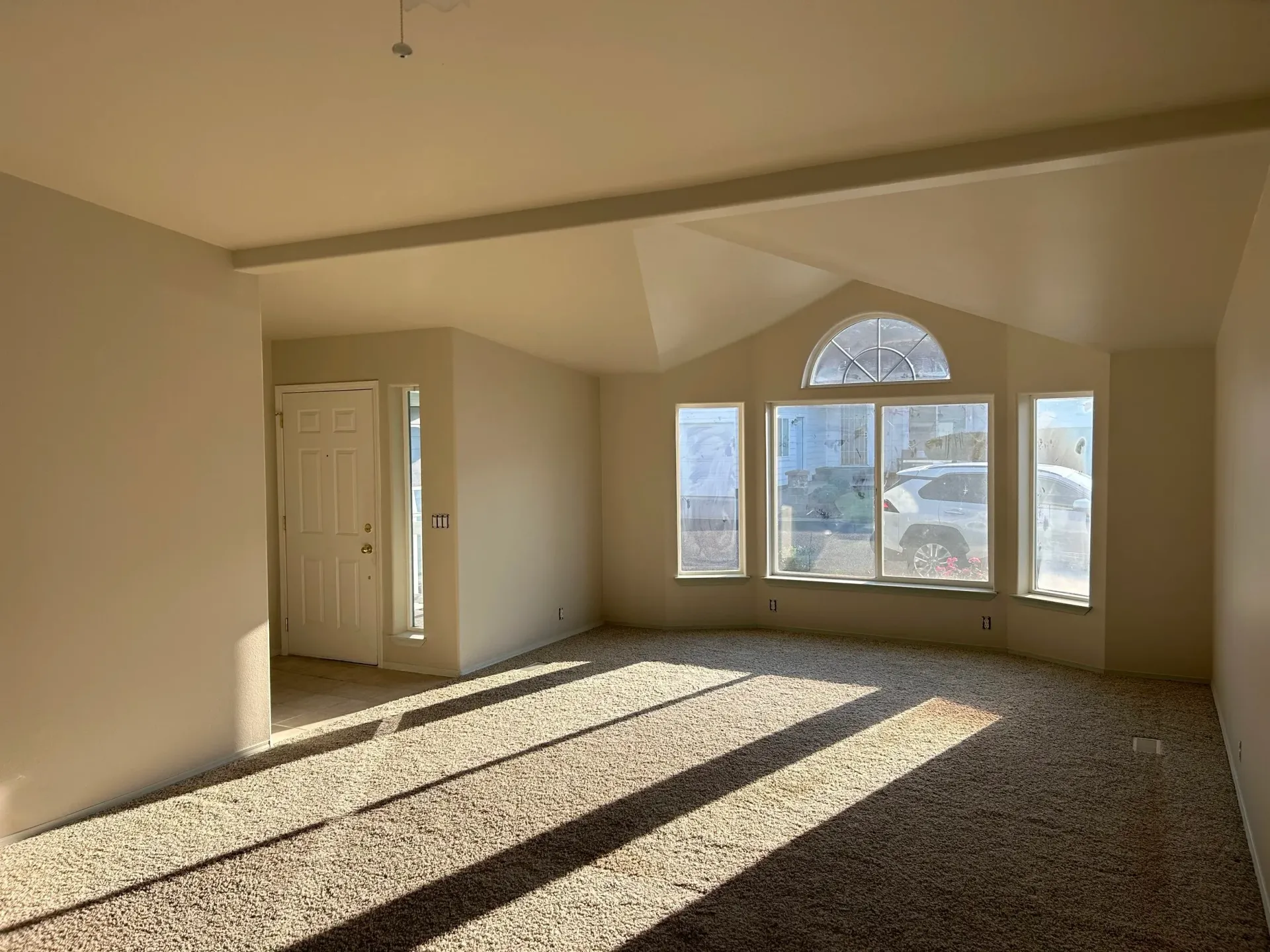 Empty living room with sunlight, arched window, and beige carpet.