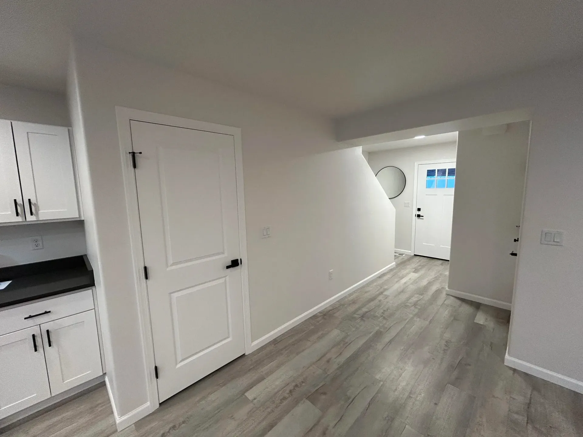 Interior view of a hallway with white doors, light gray walls, and wood-look flooring. A kitchen is visible on the left.
