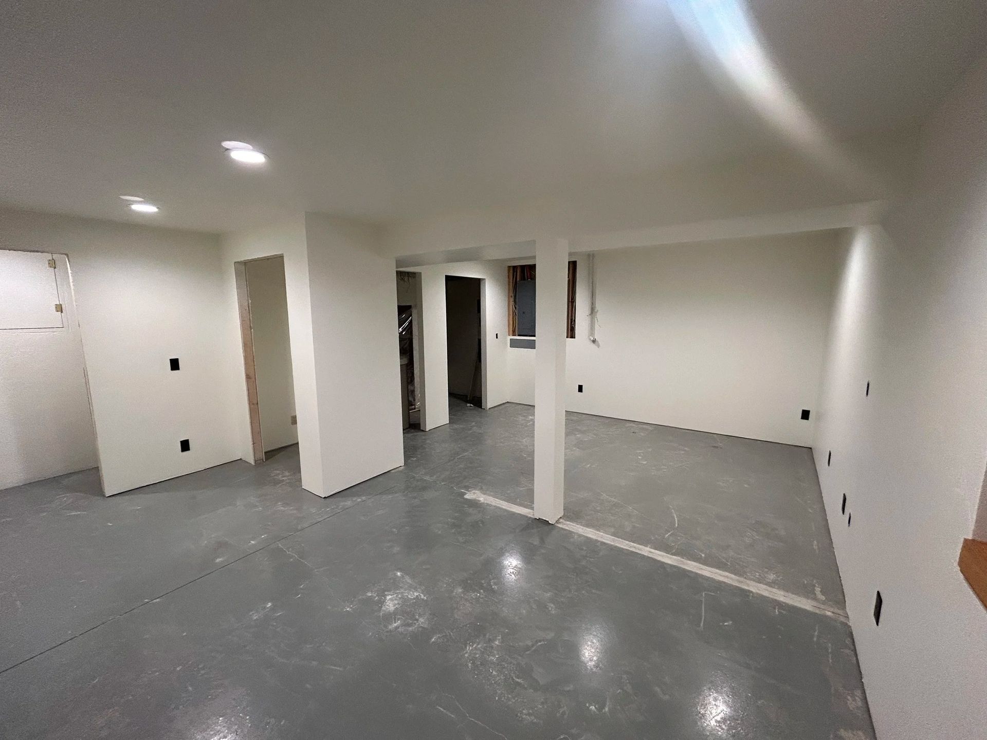 Empty basement room with white walls, gray floor, recessed lighting, and exposed electrical outlets.