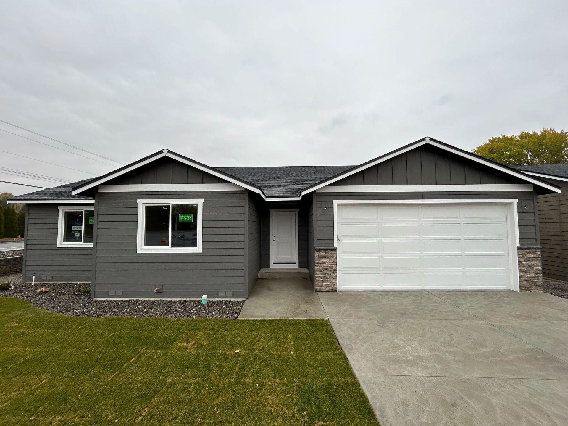 Gray house with white trim, front door, and attached garage on a concrete driveway, overcast sky.