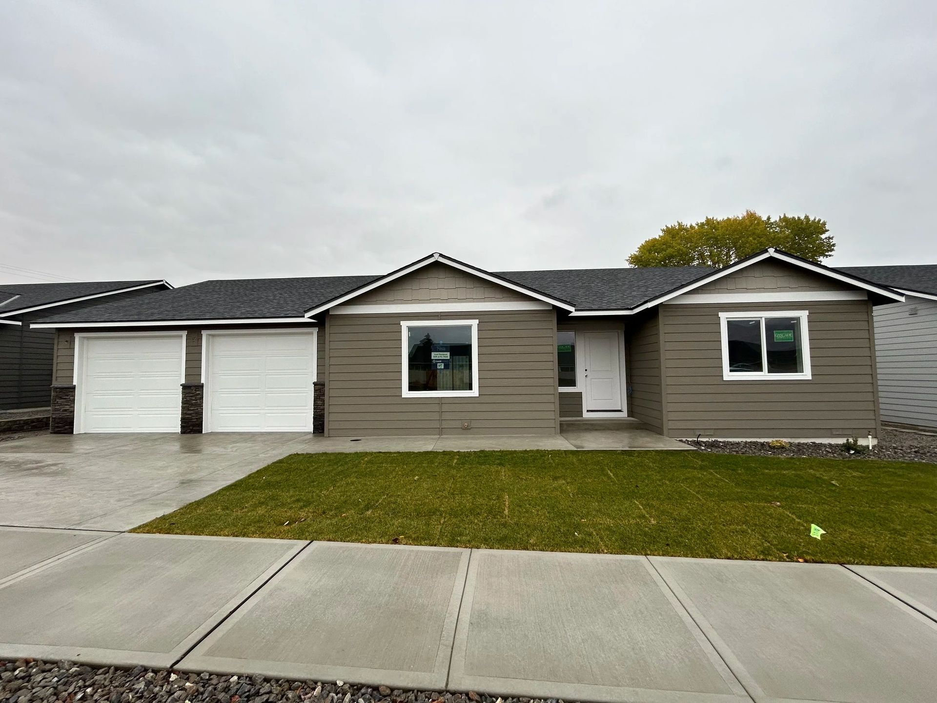 Tan house with a two-car garage, green lawn, and gray siding under a cloudy sky.