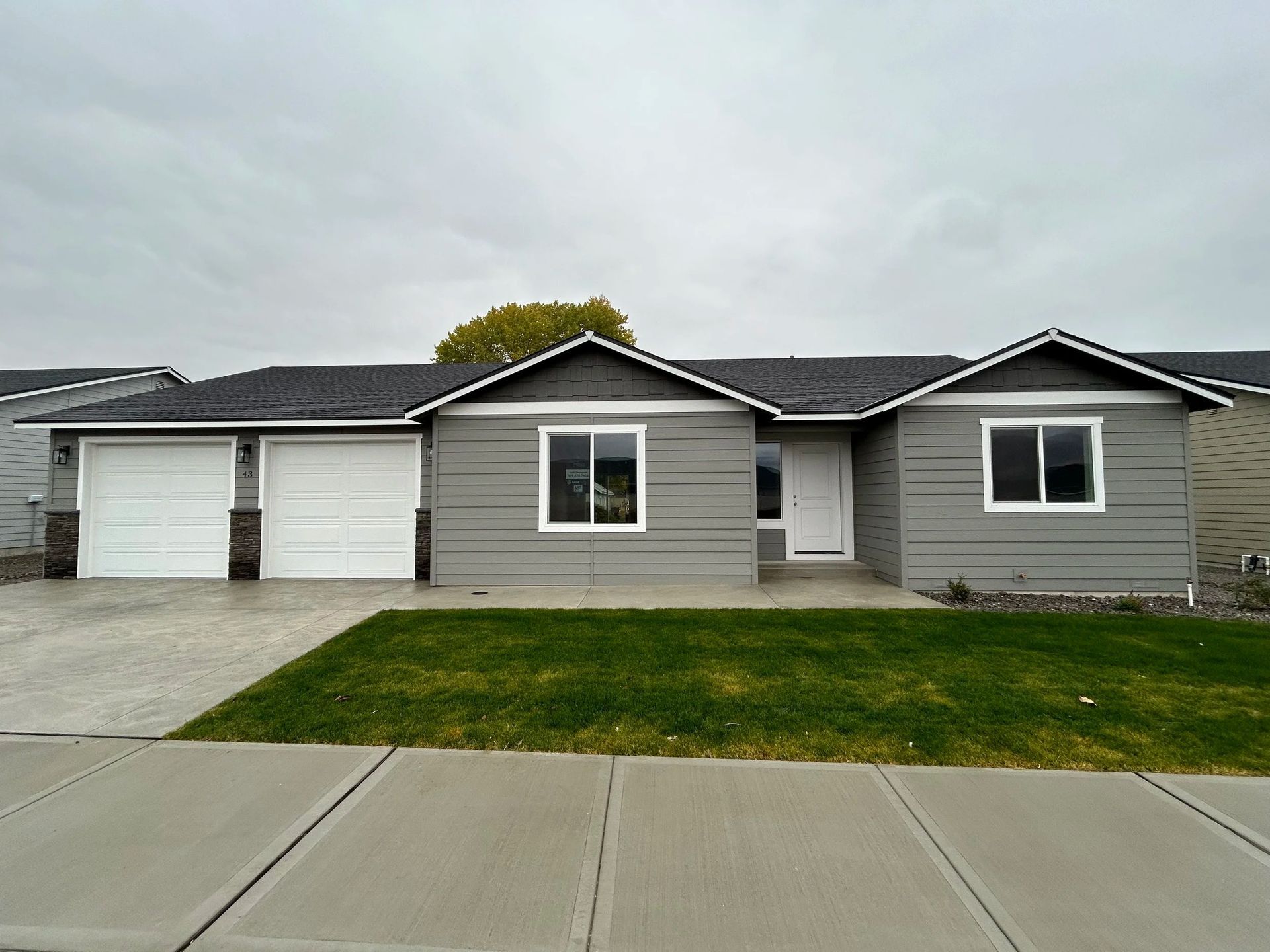 Gray house with white garage doors, lawn, and sidewalk on a cloudy day.