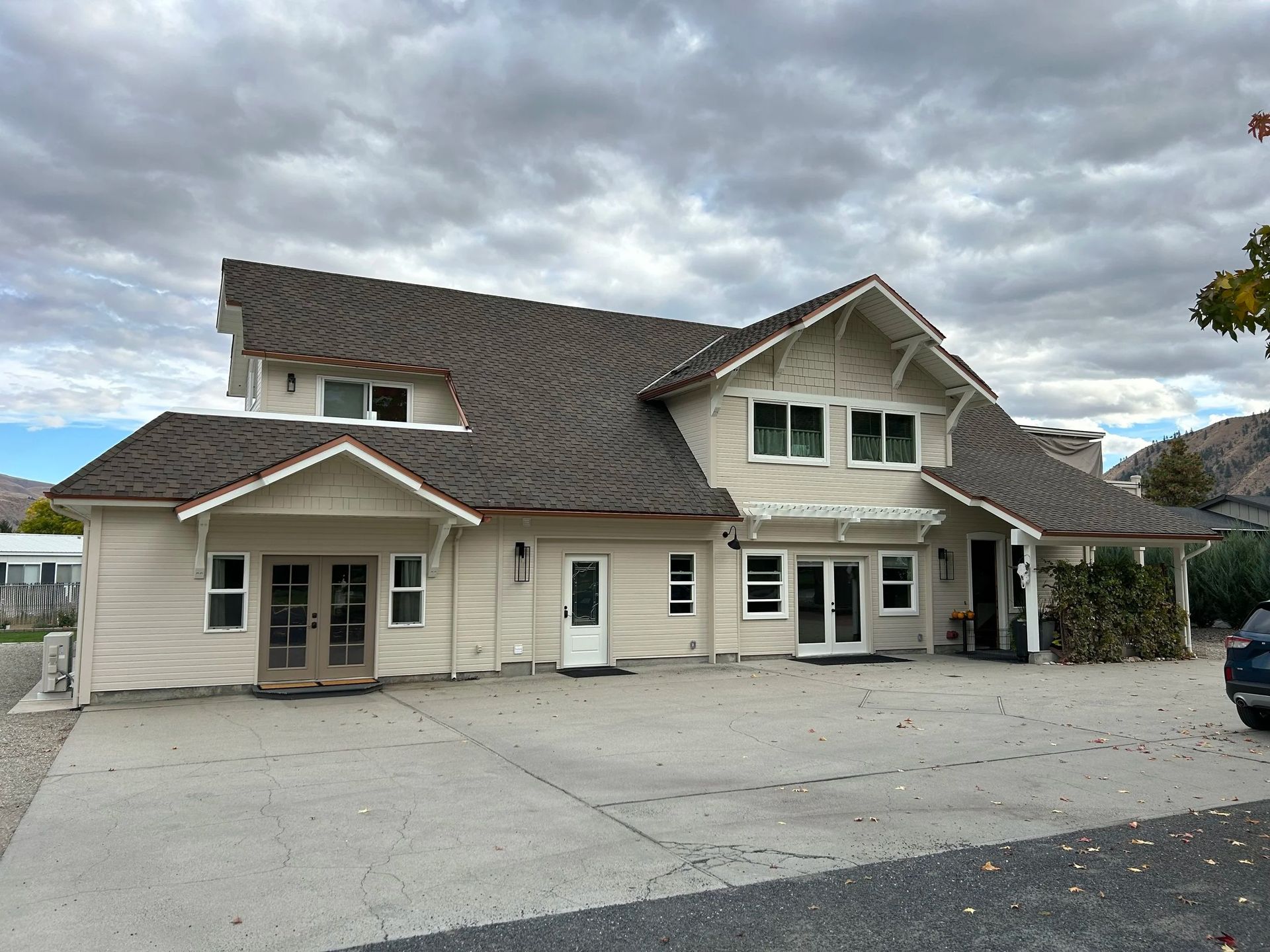Beige two-story house with dark roof and multiple windows, cloudy sky, concrete driveway.