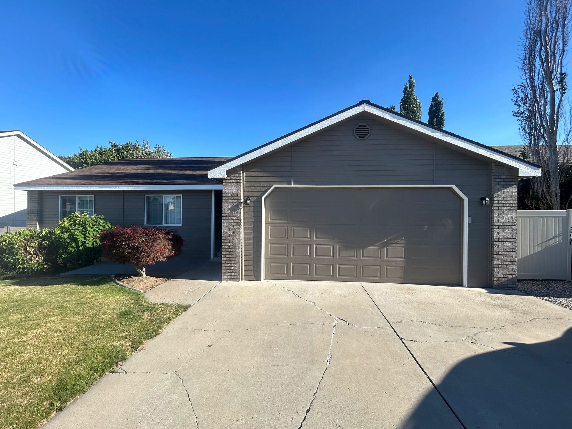 Gray house with a two-car garage, concrete driveway, and a small front yard with shrubs under a blue sky.