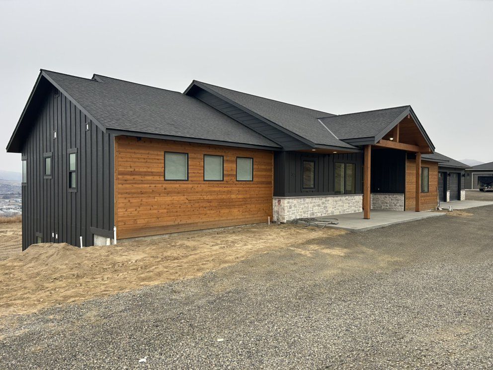 Modern one-story house with black siding, wood paneling, and a covered porch on a gravel driveway under a cloudy sky.