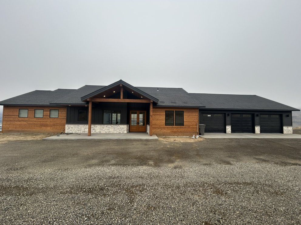 Exterior view of a modern ranch home with wood siding, a covered porch, and a three-car garage on a gravel lot.
