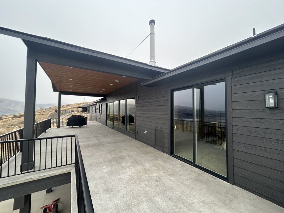 Modern home exterior with covered patio, concrete deck, and panoramic windows. Gray siding, black railing, overcast sky.