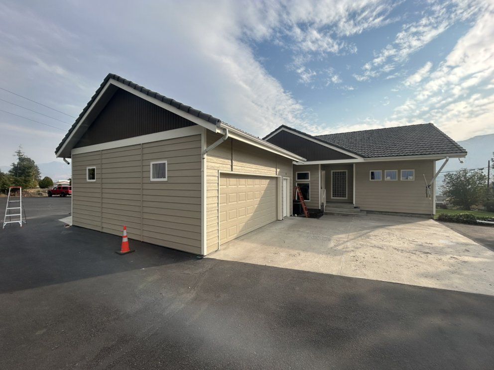 Tan house with attached garage, dark roof, and asphalt driveway under a cloudy sky.