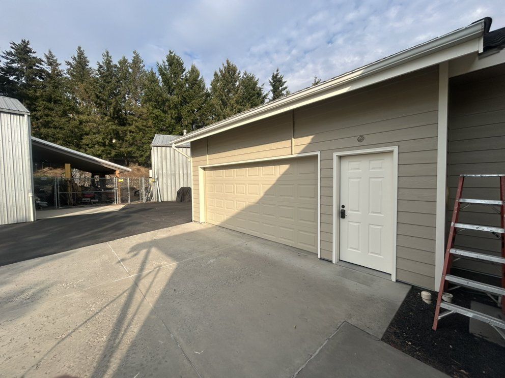 Garage and attached door, beige walls, asphalt driveway. Ladder leans against the building, with a carport visible on the left.
