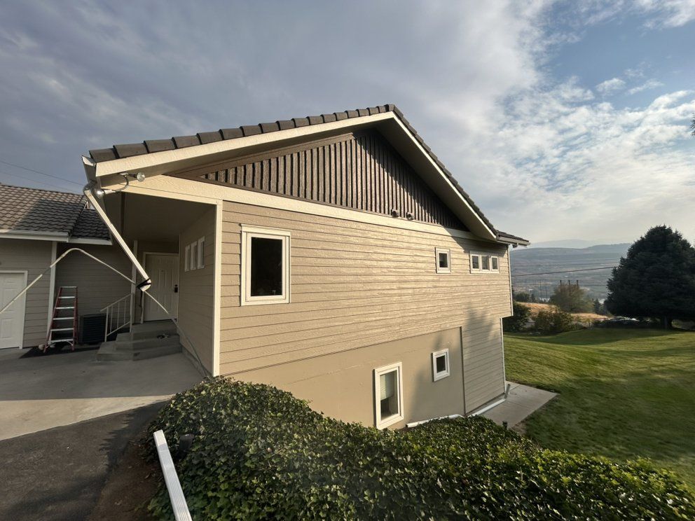 Beige house with dark roof, windows, and lush green hillside. Overcast sky.