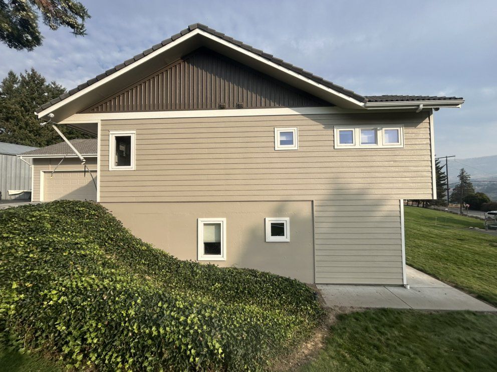 Two-story tan house with brown trim and roof, nestled into a hillside with green shrubbery.