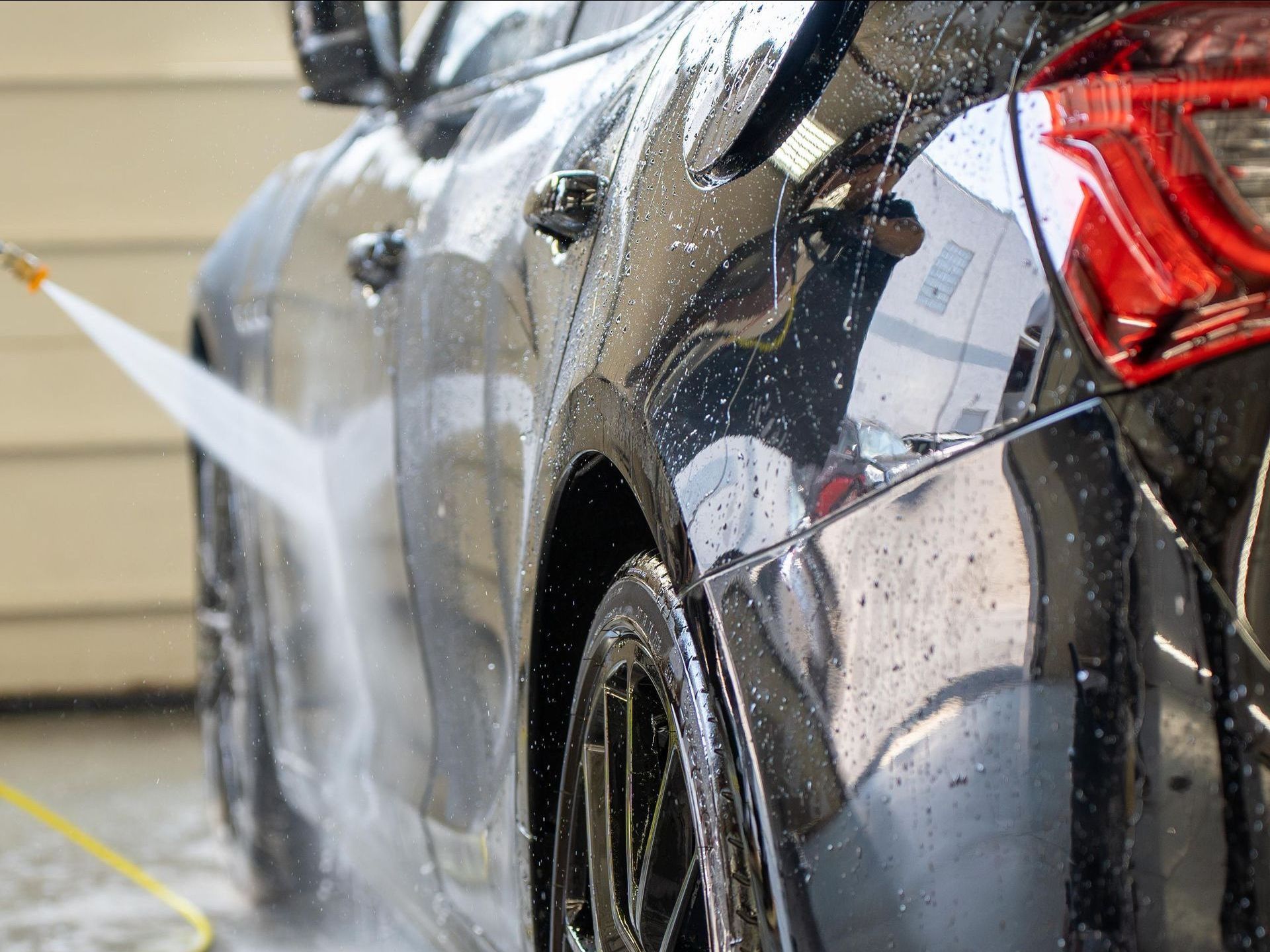 A black car being washed with a water hose, with a focus on the side and rear. Water sprays cover the car, creating glistening droplets.
