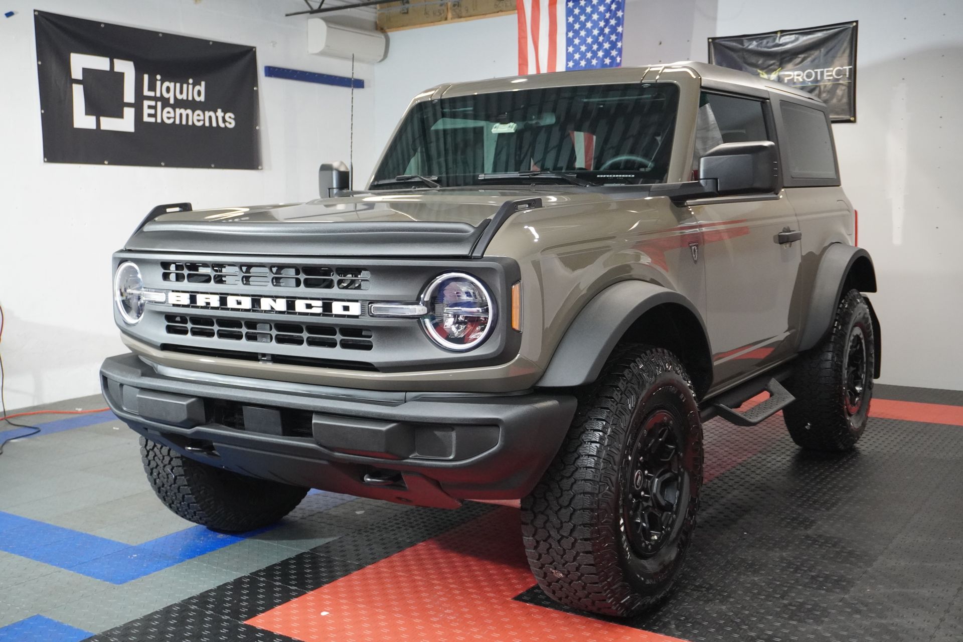 A tan Ford Bronco with black accents parked in a garage with the American flag in the background.
