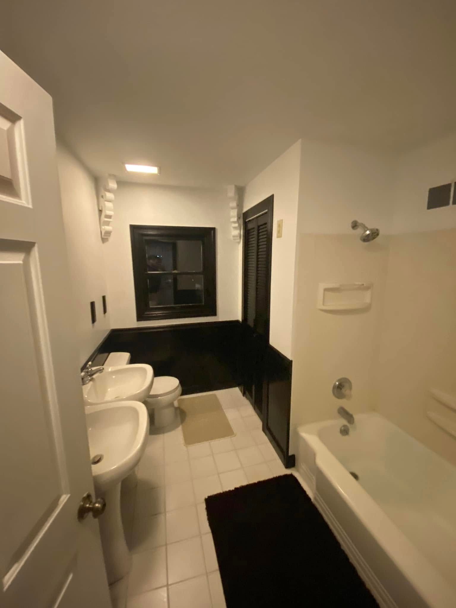A white-tiled bathroom featuring two pedestal sinks, a toilet, a shower-tub combination, and a black-framed window.