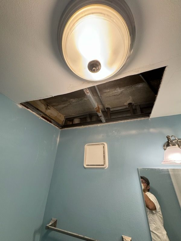 Bathroom ceiling with missing drywall panel, exposed framing, and a lit ceiling fixture above blue walls.
