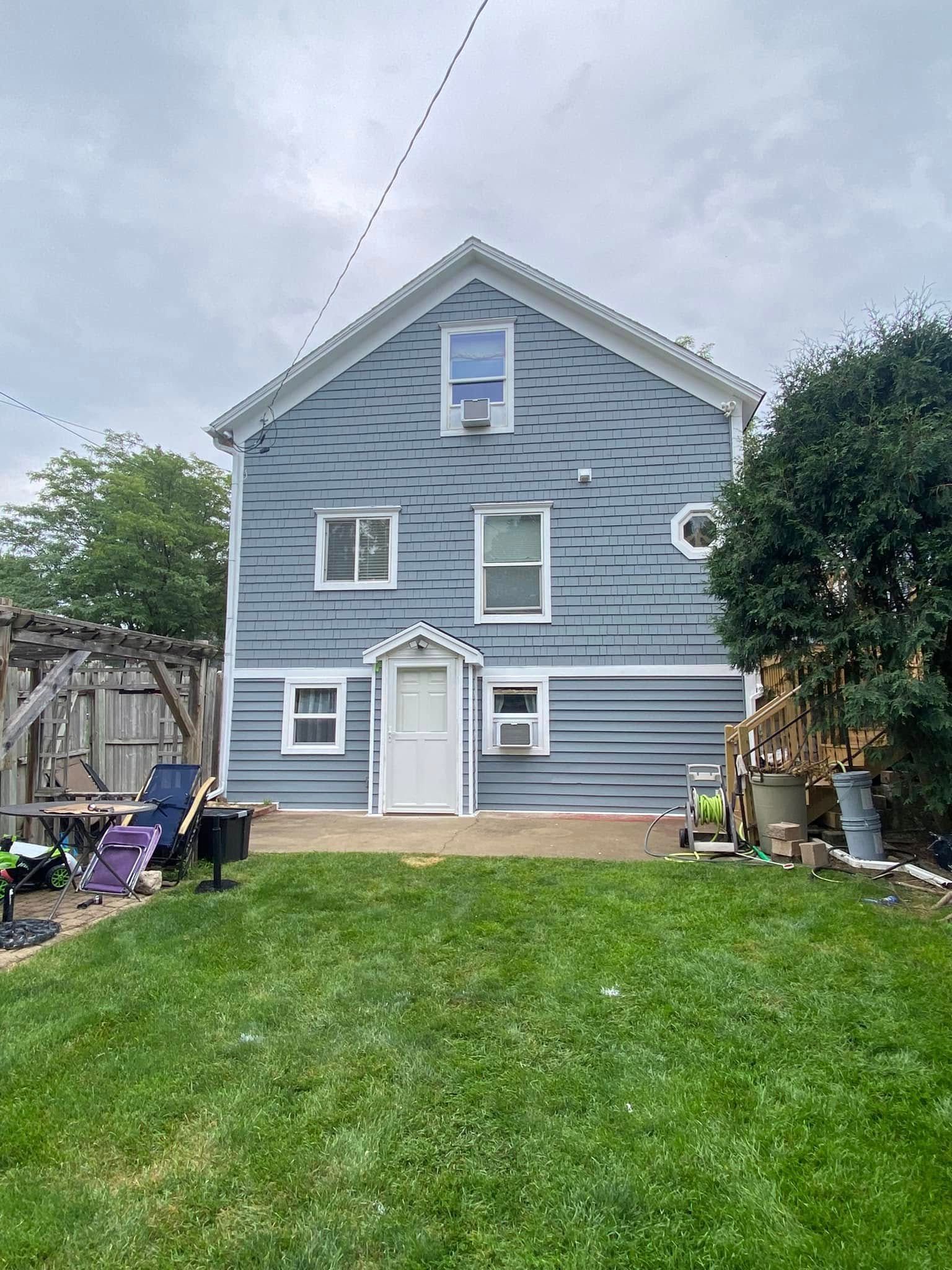 A two-story blue house viewed from a backyard, with a white door, several windows, a trellis, and a grassy lawn.
