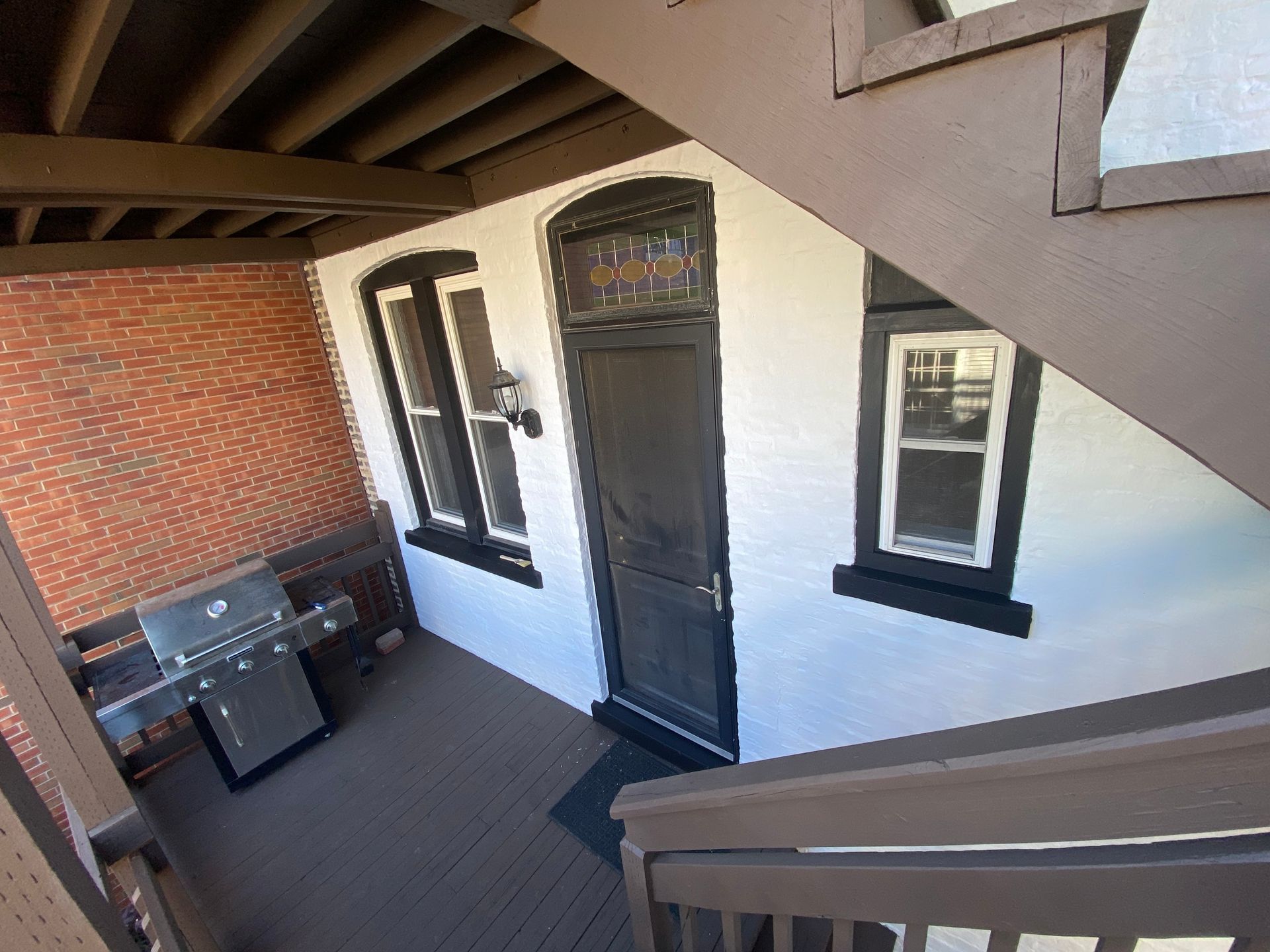 A back porch with a grill, a black door, two windows, and stairs leading up to an overhanging deck.
