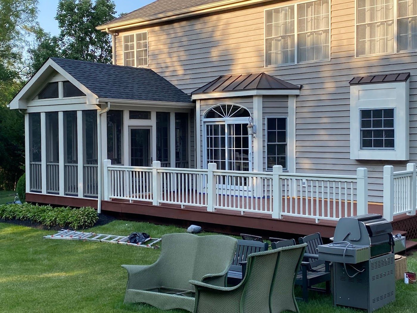 A tan house with a brown-roofed sunroom, white deck railing, and outdoor furniture on a grassy lawn.