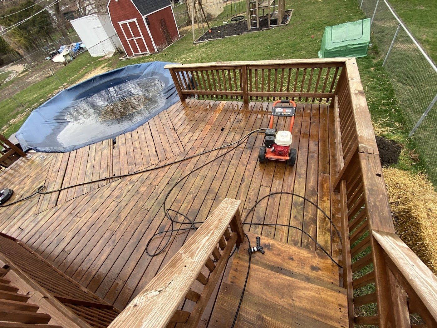 A pressure washer sits on a freshly cleaned, wet wooden deck next to an above-ground swimming pool in a backyard.