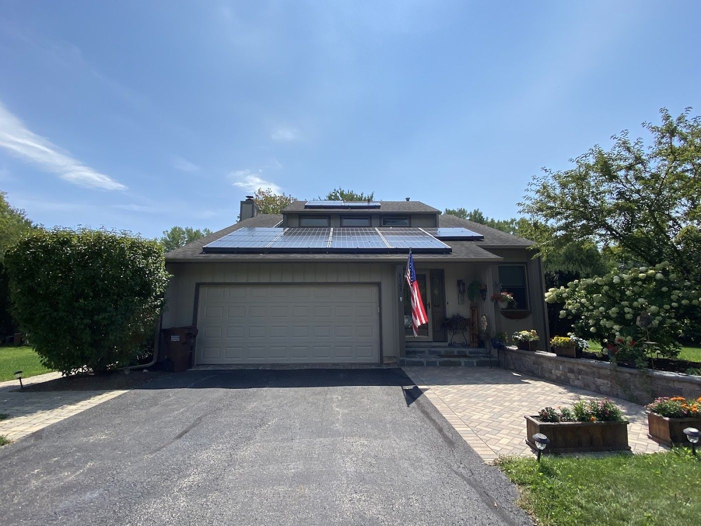 A house with a large solar panel array on the roof, a tan garage door, and a paved driveway under a clear blue sky.