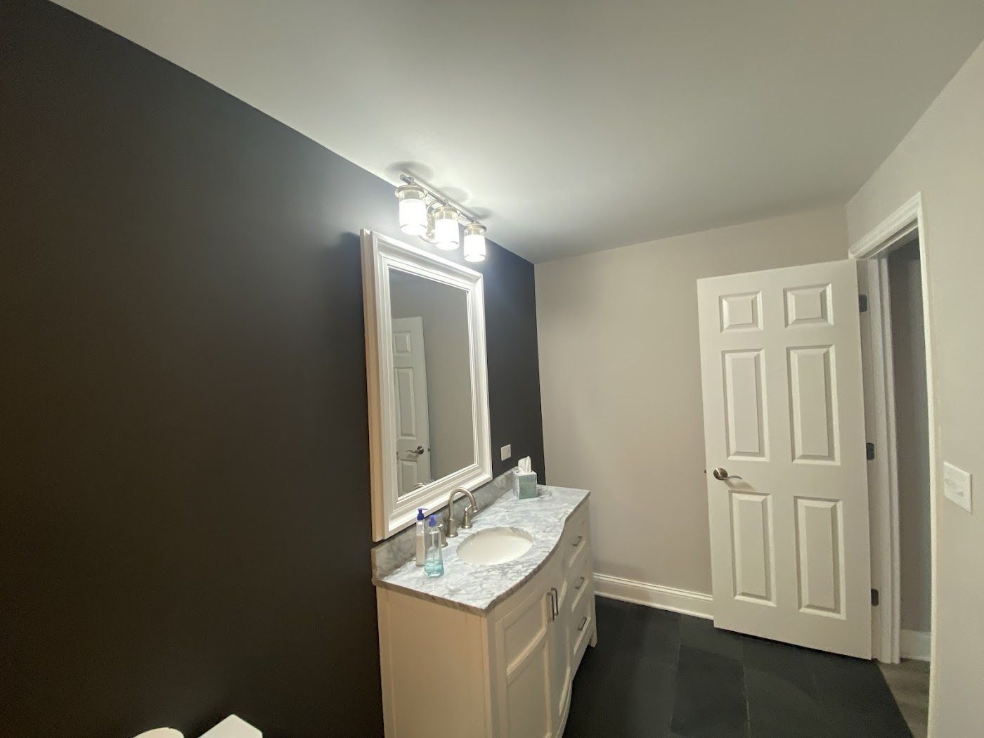 A modern bathroom with a dark brown accent wall, white vanity with marble top, light grey walls, and a white interior door.