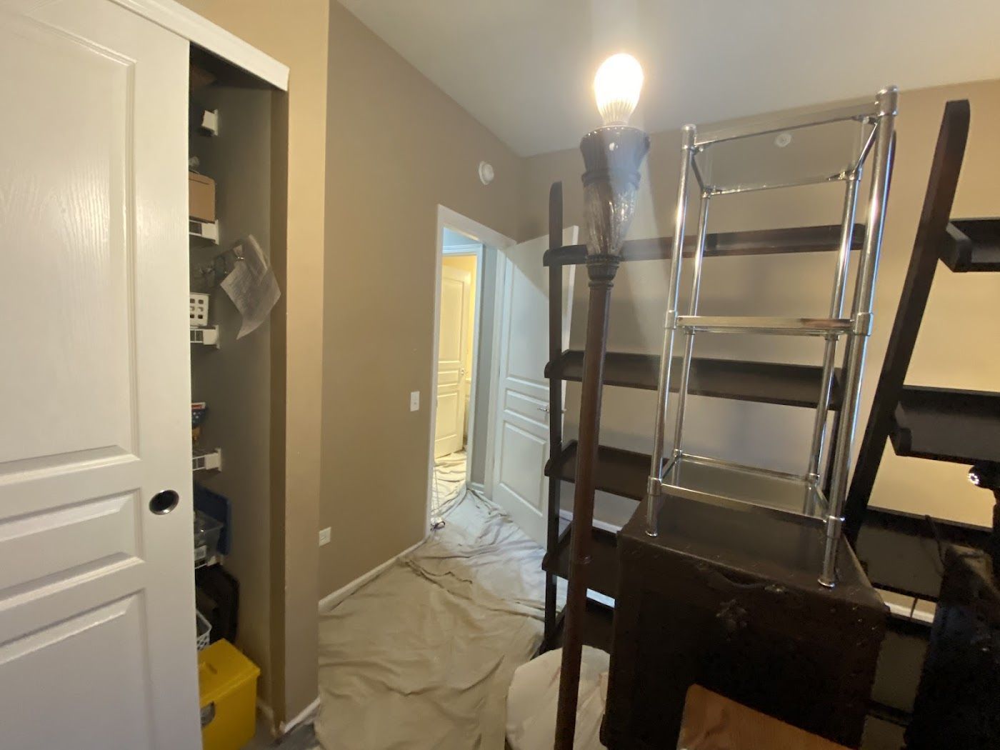 An indoor room with a closet, a metal-framed shelf, a tall lamp, and a floor covered with a drop cloth.