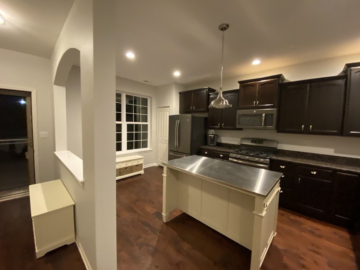 A modern kitchen with dark cabinetry, stainless steel island, and wood floors, seen from an entryway with an arched wall.
