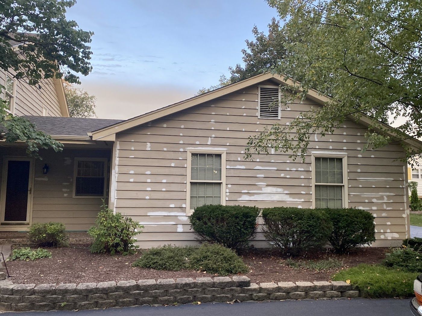 A beige, single-story house with peeling exterior siding, two windows, and a small gabled roof set behind a stone